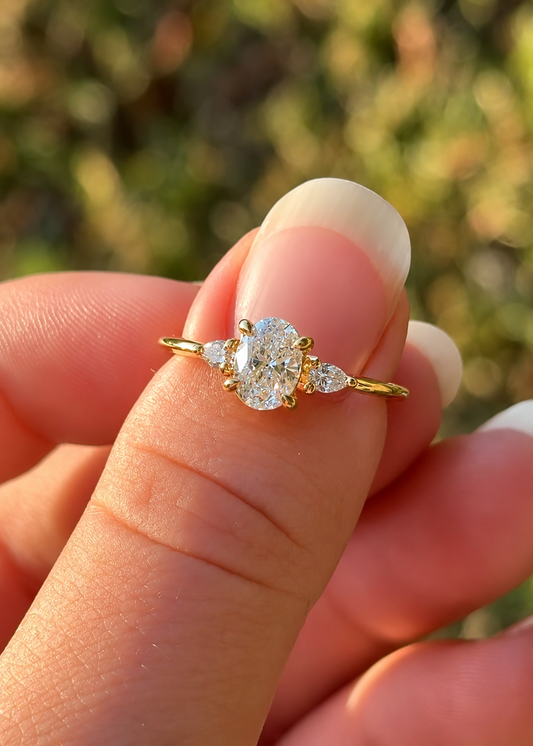 A yellow gold ring with a central oval diamond and two smaller pear-cut diamonds, held between fingers against a blurred outdoor background.